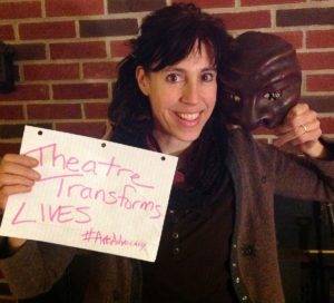 Holly holding a mask and sign that says "Theatre Transforms Lives"
