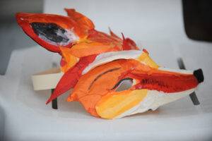 A fox mask in red, orange, white and gold. The colors are tissue paper.